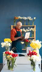 An elderly male flowers of European appearance smiles confidently while using laptop in a flower shop.