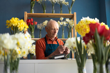 An elderly European man manager in flower shop arranges delivery using smartphone.