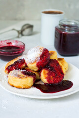 Breakfast, cheesecakes on a plate topped with berry sauce, a cup of coffee, a jar of jam..