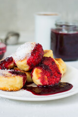 Breakfast, cheesecakes on a plate topped with berry sauce, a cup of coffee, a jar of jam..