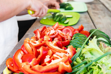Sliced red bell peppers, tomatoes, and a variety of leafy greens and hands slicing cucumbers on...