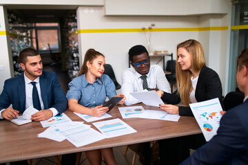 Mixed ethnicity team engages in strategic planning at boardroom table with digital tablets, financial reports. Colleagues exchange ideas in corporate meeting. Pro group focuses on graphs, data.