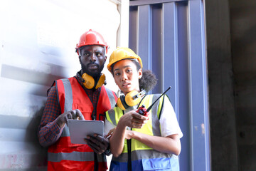 Two African workers wear safety vest and helmet at logistic shipping cargo containers yard. Happy engineer woman uses digital tablet to discuss results with colleague and pointing away at workplace.