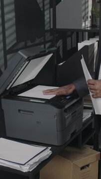 Vertical side tilt footage of male lawyer in suit making copy or scan of legal documents using multi-function printer in modern office