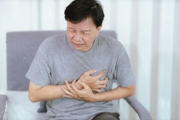 An elderly Asian man having a heart attack sits on a sofa in the living room. The elder clutched...