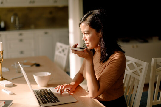 Businesswoman using laptop while talking through smart phone speaker sitting at dining table in kitchen