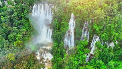 Fototapeta premium Within the depths of the dense, verdant jungle, a colossal waterfall roars with untamed vigor. The drone's lens unveils the raw, unbridled force of nature. Lush woods teeming with life. Thailand. 