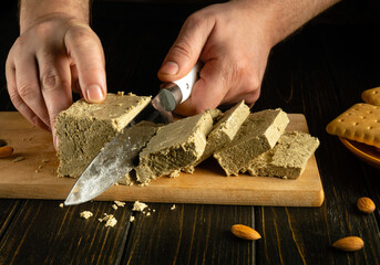 Slicing halva for dessert with a knife in the hands of a chef. A cook cuts halva on a kitchen board before preparing a sweet breakfast.