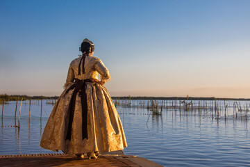 A fallera stands at the edge of the Albufera lagoon, wearing a golden dress, admiring the beauty of the landscape at sunset.