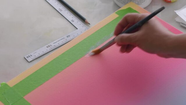 A diverse biracial female artist, paints a clear gesso onto a gradient painted artist canvas and masking tape