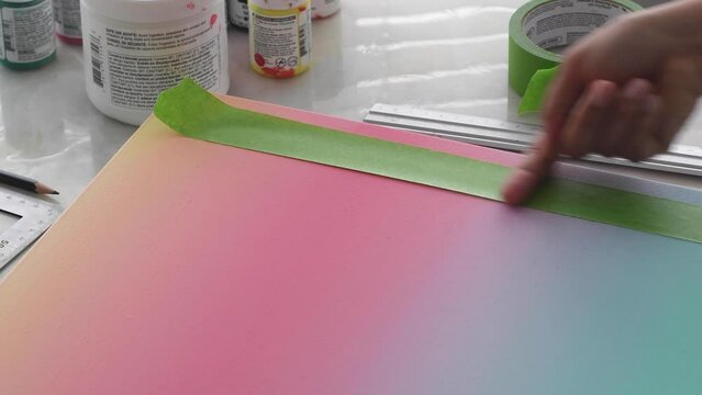 A diverse biracial female hand of an artist places masking tape on a gradient painted artist canvas.