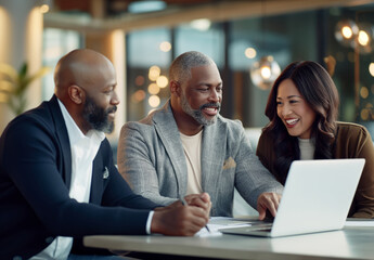 Three diverse business people, colleagues sit around an open laptop, smiling and using tablets to discuss documents for a new marketing campaign in a modern, casual office setting.