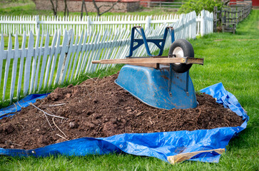 Wheelbarrow and pile of soil on a tarp in rural landscape by white wooden picket fence