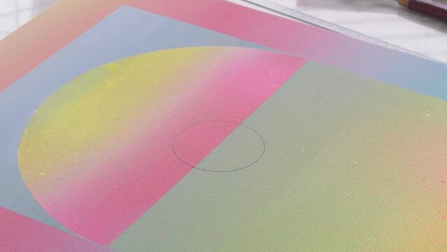 A diverse biracial female artist, uses a circle ruler and a pencil to draw out a circle on a gradient painted artist canvas.