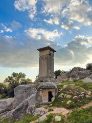A striking ancient tomb towers over the historical site of Xanthos in Kaş, Antalya, Turkey, showcasing unique architecture against a scenic sky backdrop.