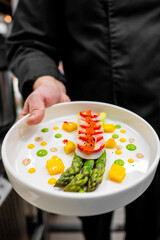 A chef in a professional kitchen presents an artfully arranged gourmet dish. The plate features green asparagus, sliced strawberries, and colorful sauces. The chef wears a black jacket