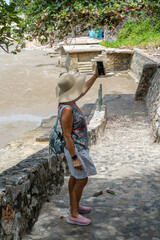 A lady in a large hat with a backpack on her back stands in the shade of a tropical tree on the stone steps of the stairs leading up the hill, taking a selfie on her phone.