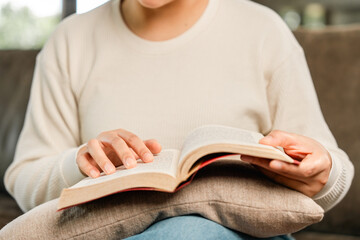 Asian woman sitting reading book on cozey couch sofa in living room with sun light morning. People stay home part time relax at home. Good time quality life rest calm happiness