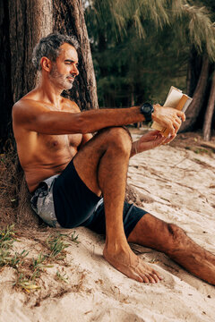 Shirtless mature man reading book while sitting near tree trunk at beach