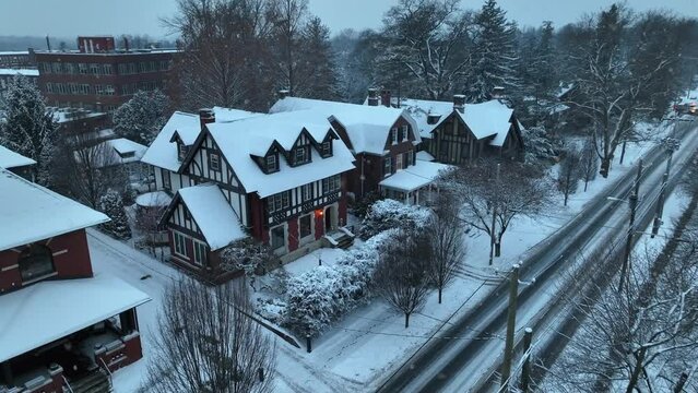 Snowy Tudor-style houses with steep gabled roofs, situated along a snow-lined street with bare trees. Aerial during snow flurries in American city at dusk.