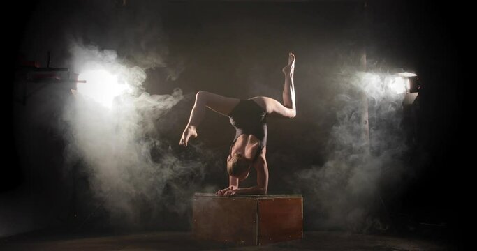 Bendy contortionist doing scorpion pose on box between spotlights and smoke