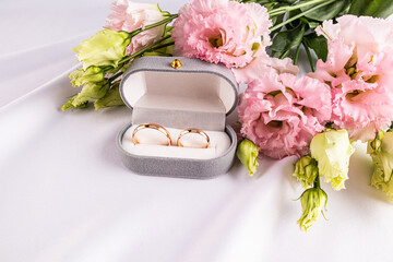 Two gold wedding classic rings in a gray velvet box on a white satin fabric with fresh pink flowers. Wedding concept. Front view
