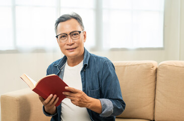 Mature senior Asian old man sitting reading book on cozey couch sofa in living room with sun light morning. People old man stay home part time relax at home reading book.