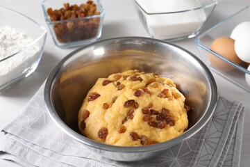 Dough with raisins in bowl and ingredients on white table, closeup