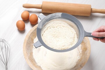 Woman sieving flour into bowl at white wooden table, above view