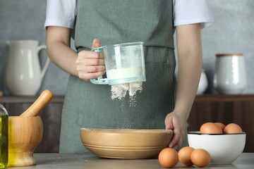 Woman sieving flour into bowl at table in kitchen, closeup