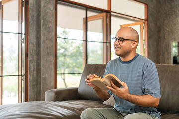 Asian man sitting reading book on cozey couch sofa in living room with sun light morning. People...