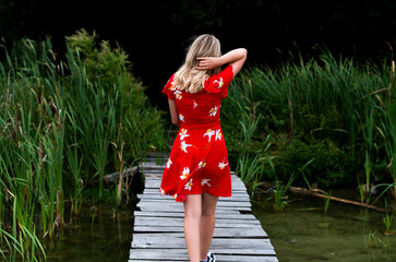 Dziewczyna w czerwonej sukience idąca pomostem na tle zielonej roślinności jeziora 