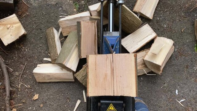 A hydraulic horizontal log splitter is splitting the wood log of an ash tree for burning by driving a wedge. The wood splitter is surrounded by a pile of logs already split.