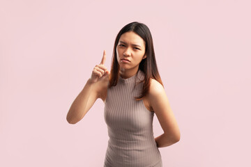 A young Asian woman pointing upward with upset facial expression, wearing a nude brown shirt on a pink background with empty copy space for advertising content