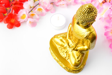 A golden statue of a Buddhist figure meditating decorate with colorful flowers and round white candle isolated on white background. Concept for Vesak Day and Enlightenment Day