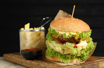 Burger with delicious patty, french fries and sauce on wooden table against dark background