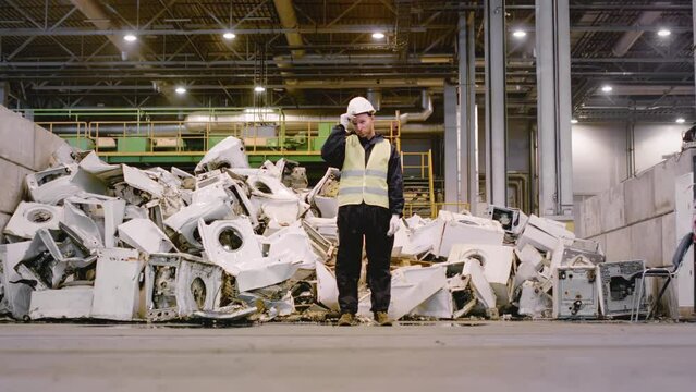 Man in helmet stands against pile of broken washing machine bodies at recycling plant. Professional worker presence exudes sense of authority and expertise overseeing operations in factory