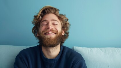 Smiling Caucasian young man with a beard sits on the sofa with his eyes closed and dreams. Blue background with copy space.