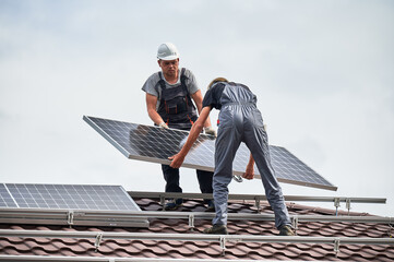 Men technicians carrying photovoltaic solar modul on roof of house. Workmen in helmets installing solar panel system outdoors. Concept of alternative and renewable energy.