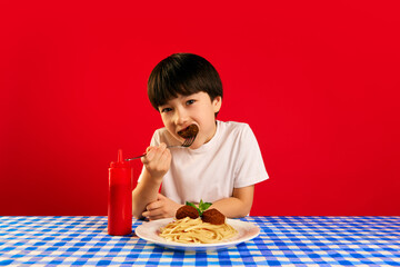Young Korean boy enjoying plate of spaghetti and meatballs at table with blue and white checked tablecloth against red background. Concept of food, childhood, emotions, meal, menu, pop art