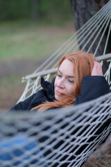 Portrait of a woman. The woman is in a hammock. The woman is wearing a black windbreaker. The woman has red hair.