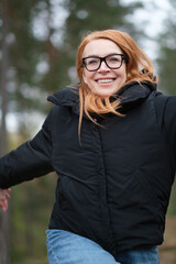 A smiling woman runs through the forest. The woman is wearing a black windbreaker, blue jeans, black glasses.