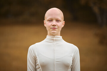 Portrait of young hairless girl with alopecia in white cloth on autumn park background, bald pretty...