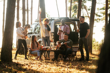 Near the car that is parked behind them. Group of friends are having fun together in the forest