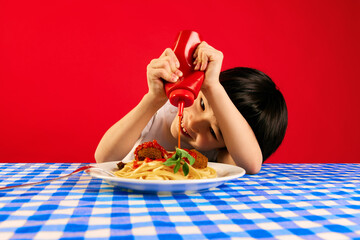 Playful kid, little boy sitting at table and squeezing ketchup sauce on plate with spaghetti and meatballs against red background. Concept of food, childhood, emotions, meal, menu, pop art