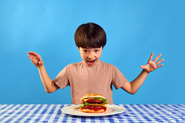 Little boy, child looking at table with delicious home made burger with happy and excited face on blue background. Delicious burger with meat and cheese. Concept of food, childhood, emotions, pop art