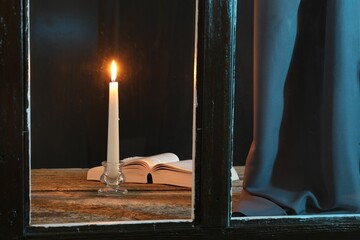Burning candle and Bible on wooden table at night, view through window