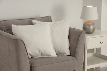 Soft white pillows on armchair, bedside table and lamp near light wall indoors