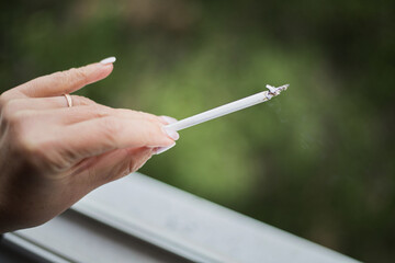 A woman’s hand holding a lit cigarette, female smoking, smoking thin cigarettes, women’s cigarettes, women’s health and tobacco, a cigarette in a woman’s hand