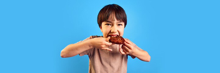 Little boy, child emotionally biting fried meat, cutlet from burger against blue background....